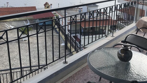 A balcony with a table and chairs, a view of the sea, and a building with a red-tiled roof. - Natalie Apartments (Sithonia)
