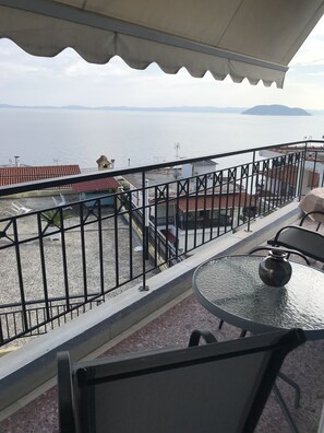 A balcony with a table and chairs, a view of the sea, and a building with a red-tiled roof. - Natalie Apartments (Sithonia)