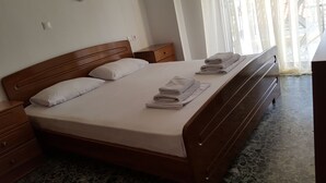 A neatly made bed with pillows and folded towels, a wooden nightstand, a dresser, and a window with sheer curtains. - Natalie Apartments (Sithonia)