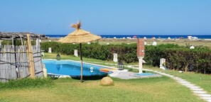 Uma área de piscina com guarda-sol de palha, um caminho de pedra e vista para a praia.