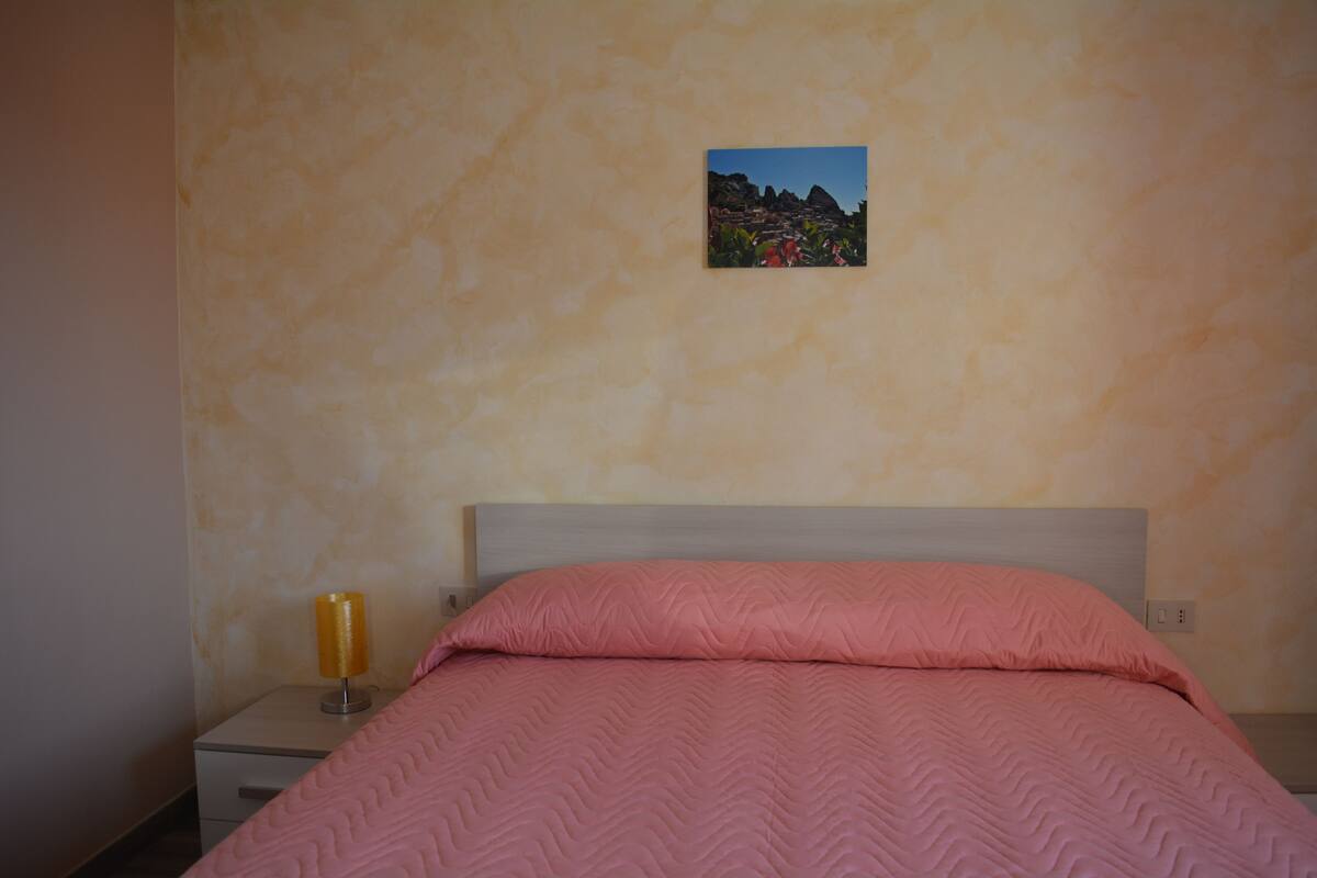A bedroom with a bed, a nightstand, a lamp, and a wall-mounted picture.