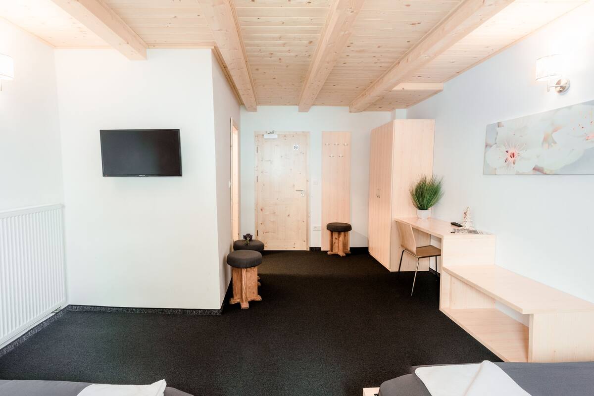 A modern hotel room with a wooden ceiling, a flat-screen TV mounted on the wall, a desk with a chair, and a large window.