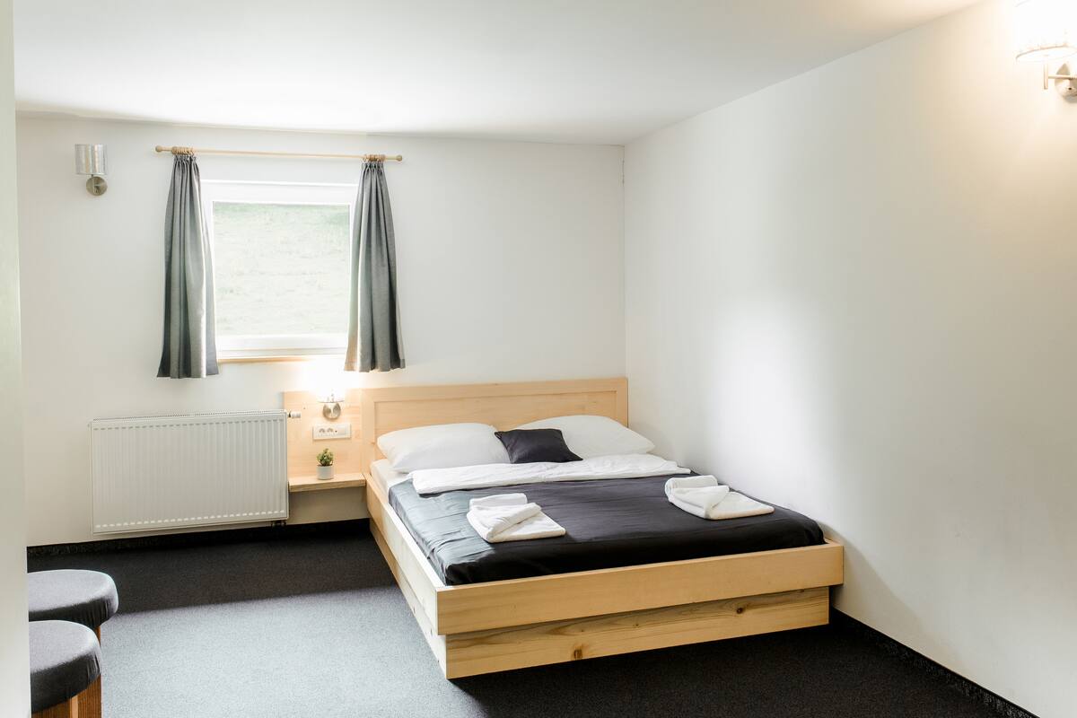 A single bed with a wooden frame, a bedside table with a lamp, a window with curtains, and a radiator.