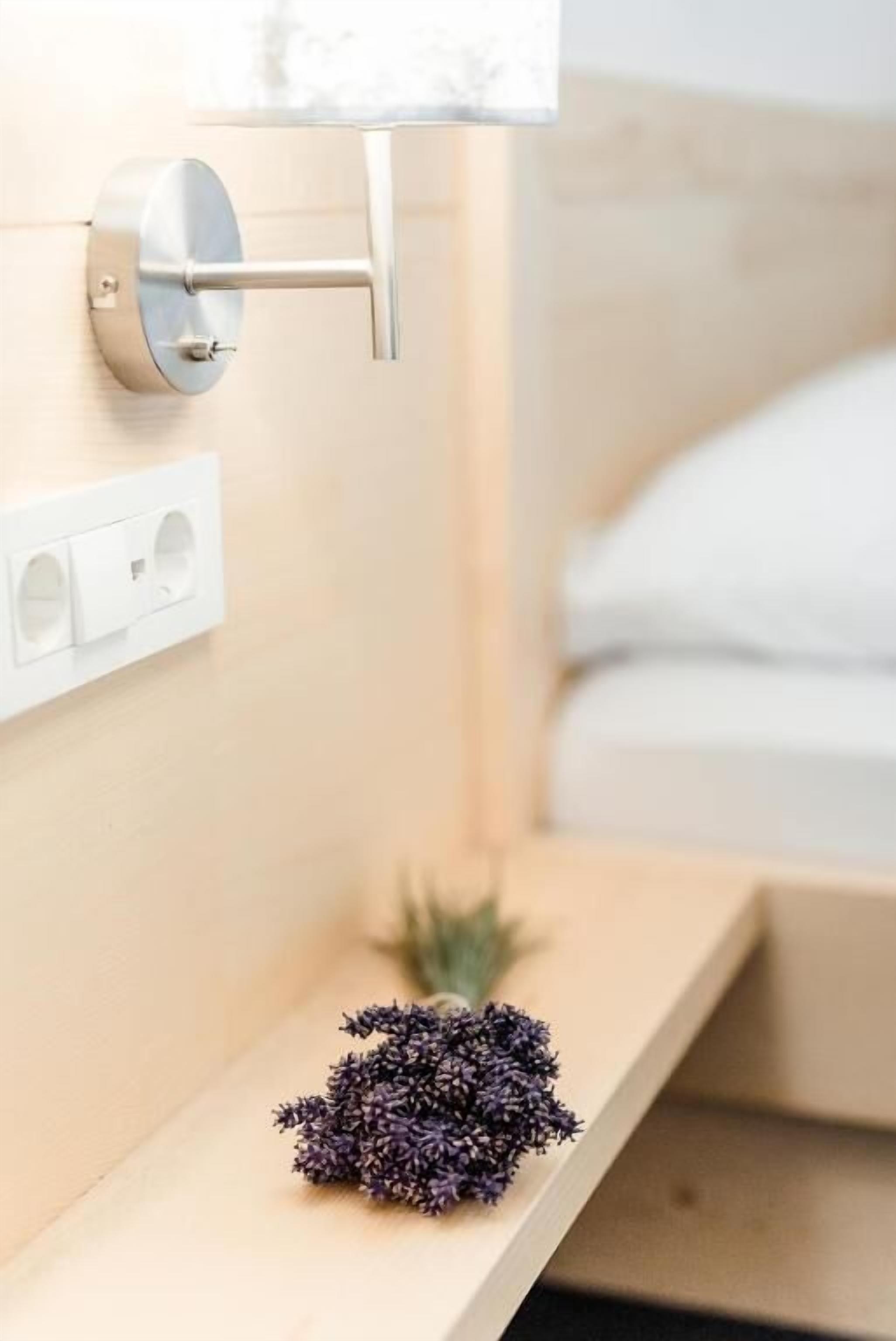 A modern bathroom with a wall-mounted toilet, a power outlet, and a small shelf with a lavender bouquet.