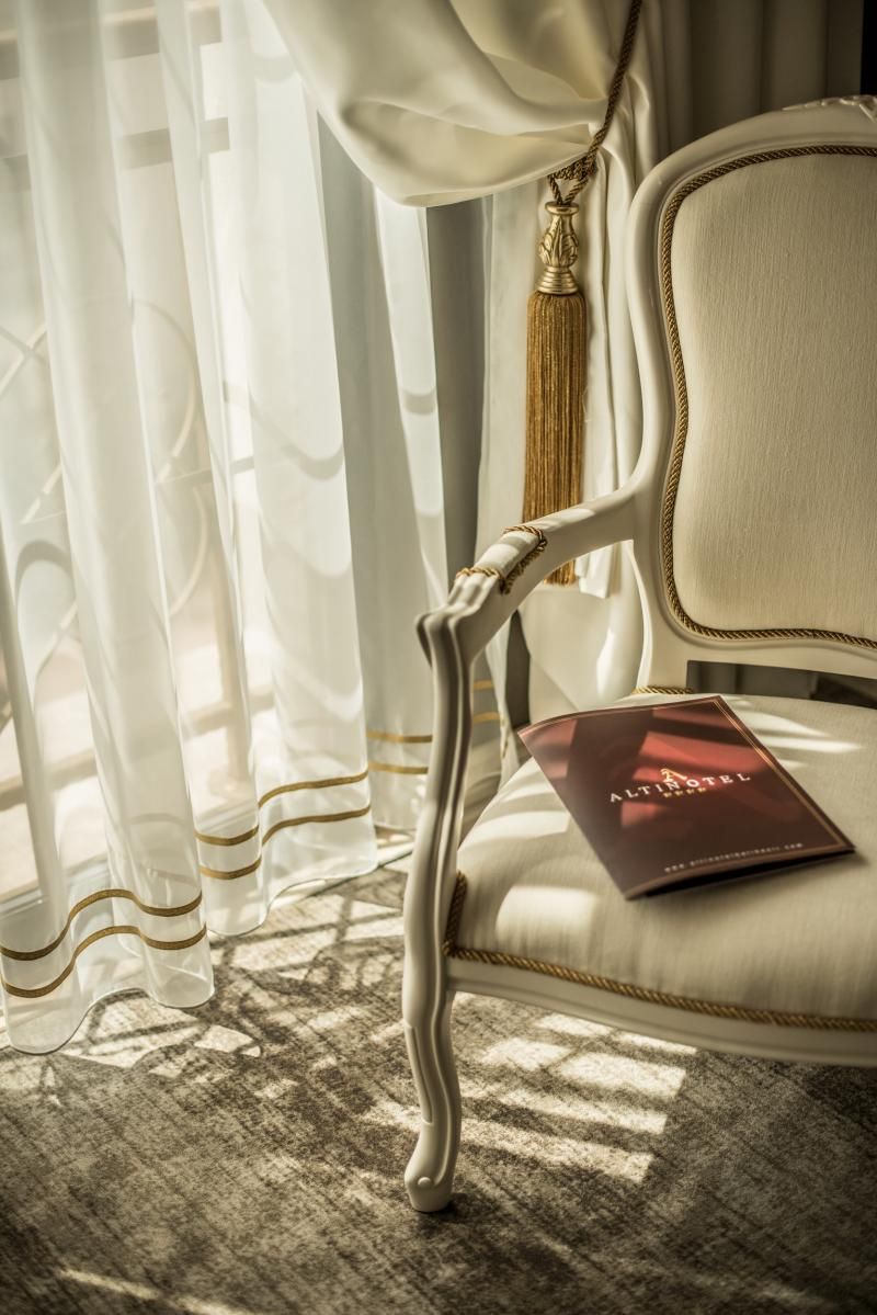 Una silla tapizada de blanco con un fleco dorado, ubicada cerca de una ventana con cortinas blancas translúcidas. Un folleto rojo con el título 'ALTINHOTEL' está colocado sobre la silla.