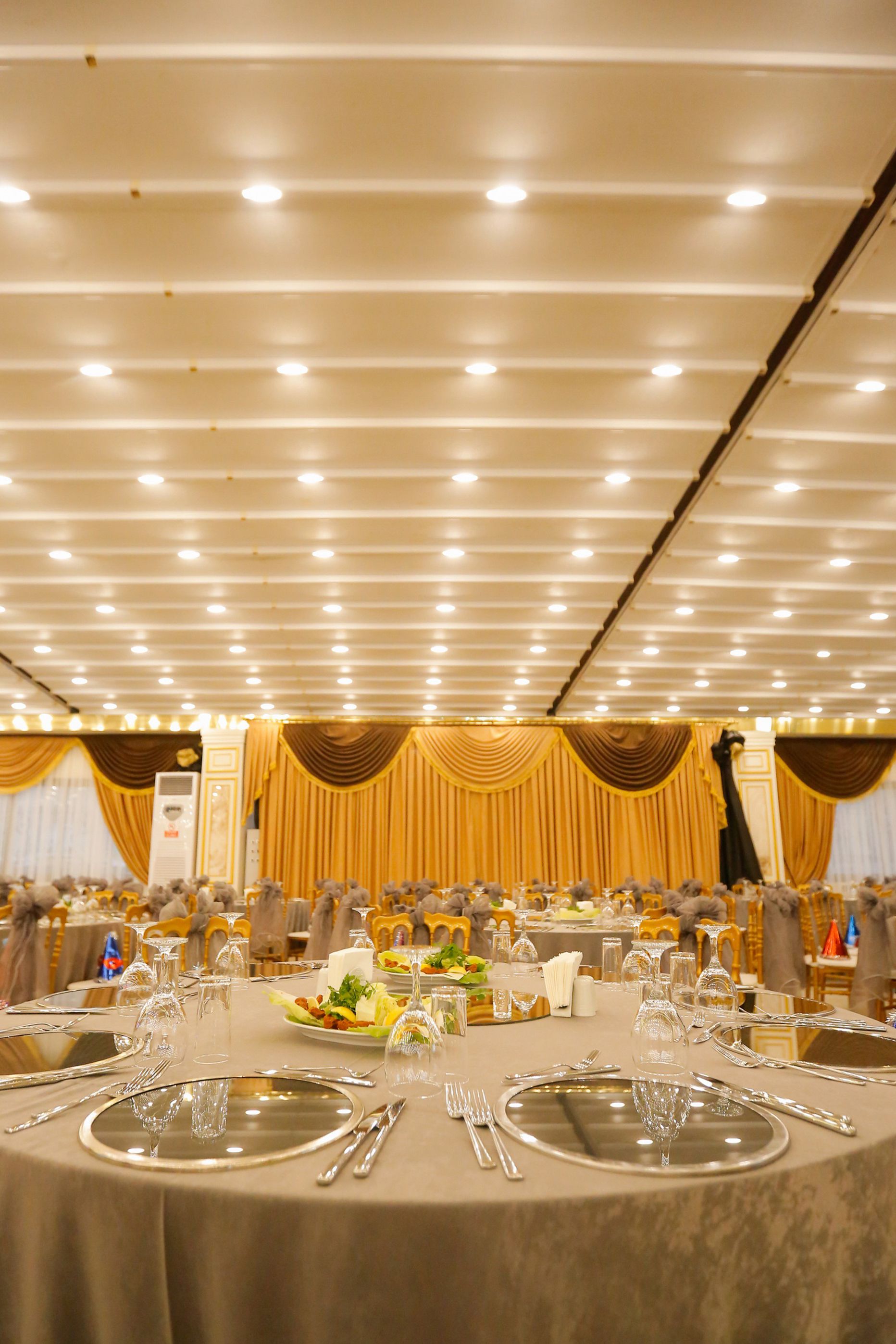 A banquet hall with round tables set for a formal event, featuring chandeliers and draped curtains.