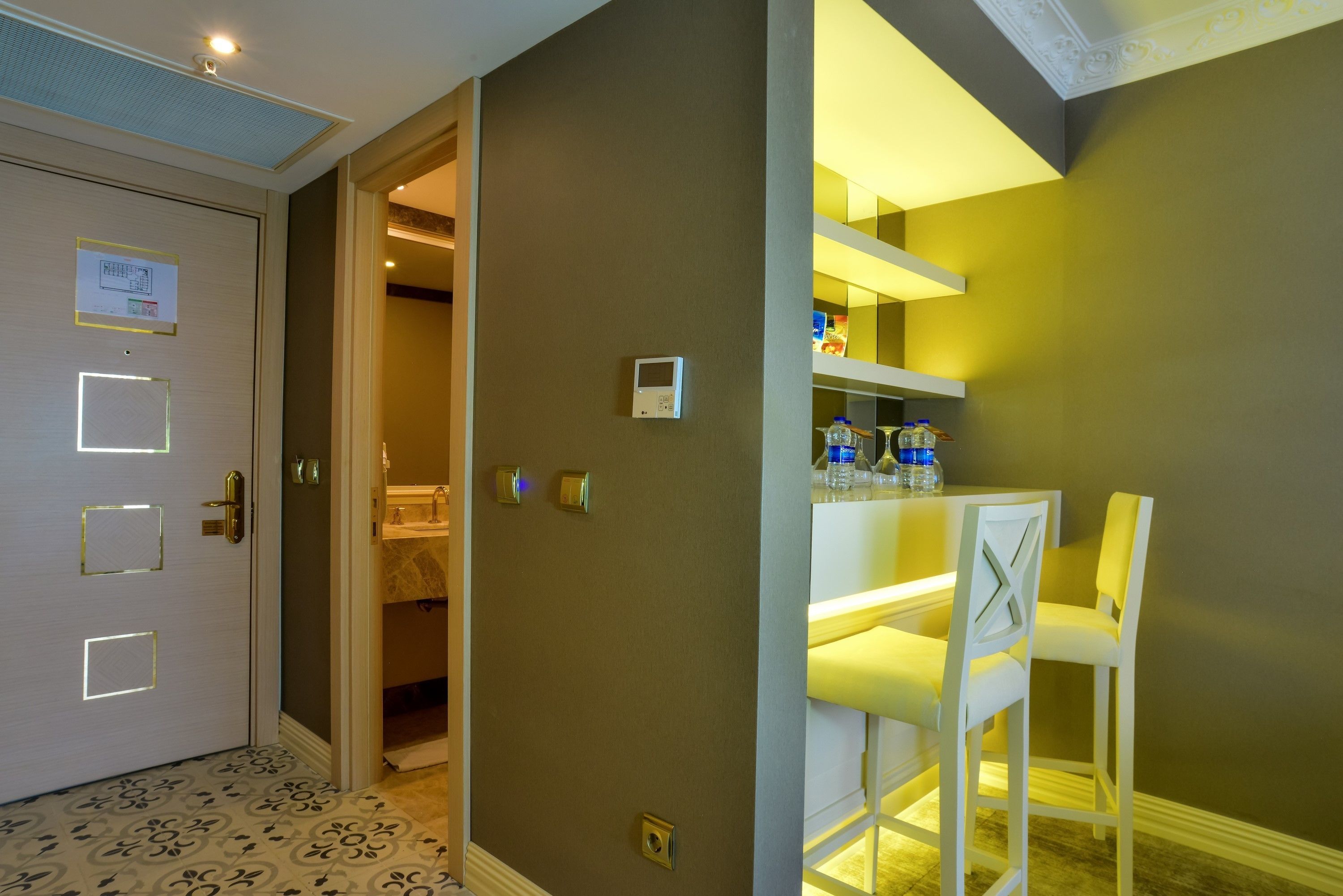 A hotel room with a closed door, a small table with chairs, a shelf with bottles, and a light fixture on the ceiling.
