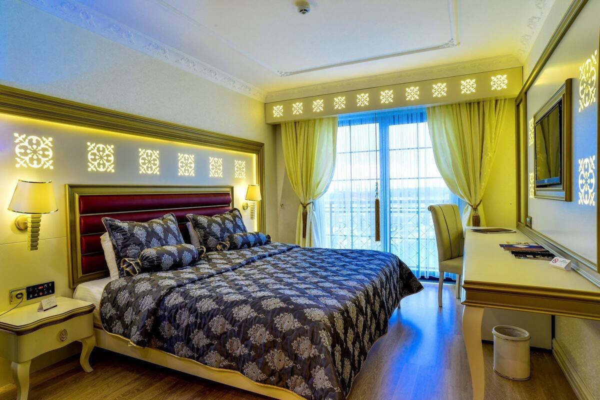 Habitación de hotel con cama, mesita de noche, lámpara, escritorio, silla y vistas al exterior.