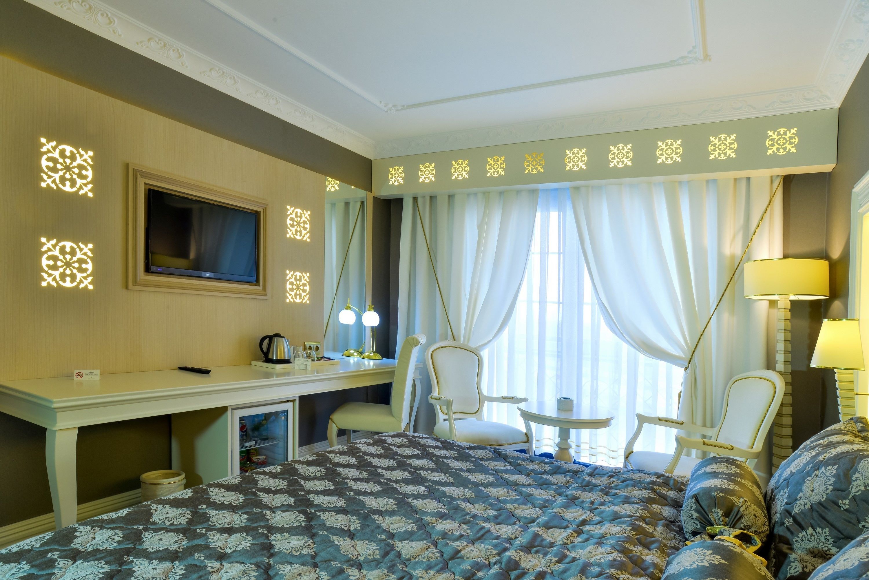 A hotel room with a bed, a desk with a TV, two chairs, a small table, and a lamp.