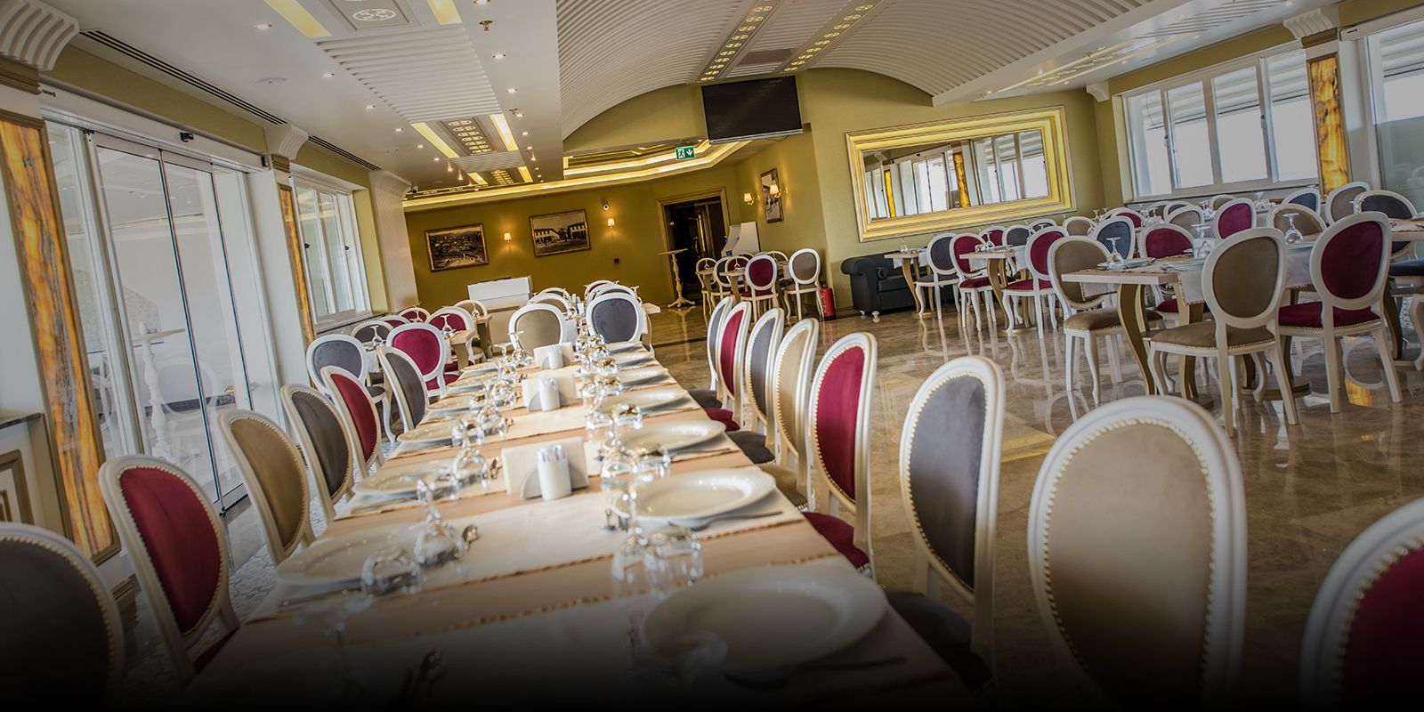 Une salle à manger élégante, avec de longues tables dressées pour accueillir de nombreux convives, des chaises dotées de coussins rouges et un grand miroir au mur.