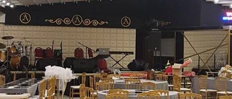 Un salón de banquetes con mesas y sillas redondas, un escenario con instrumentos musicales e iluminación empotrada en el techo.