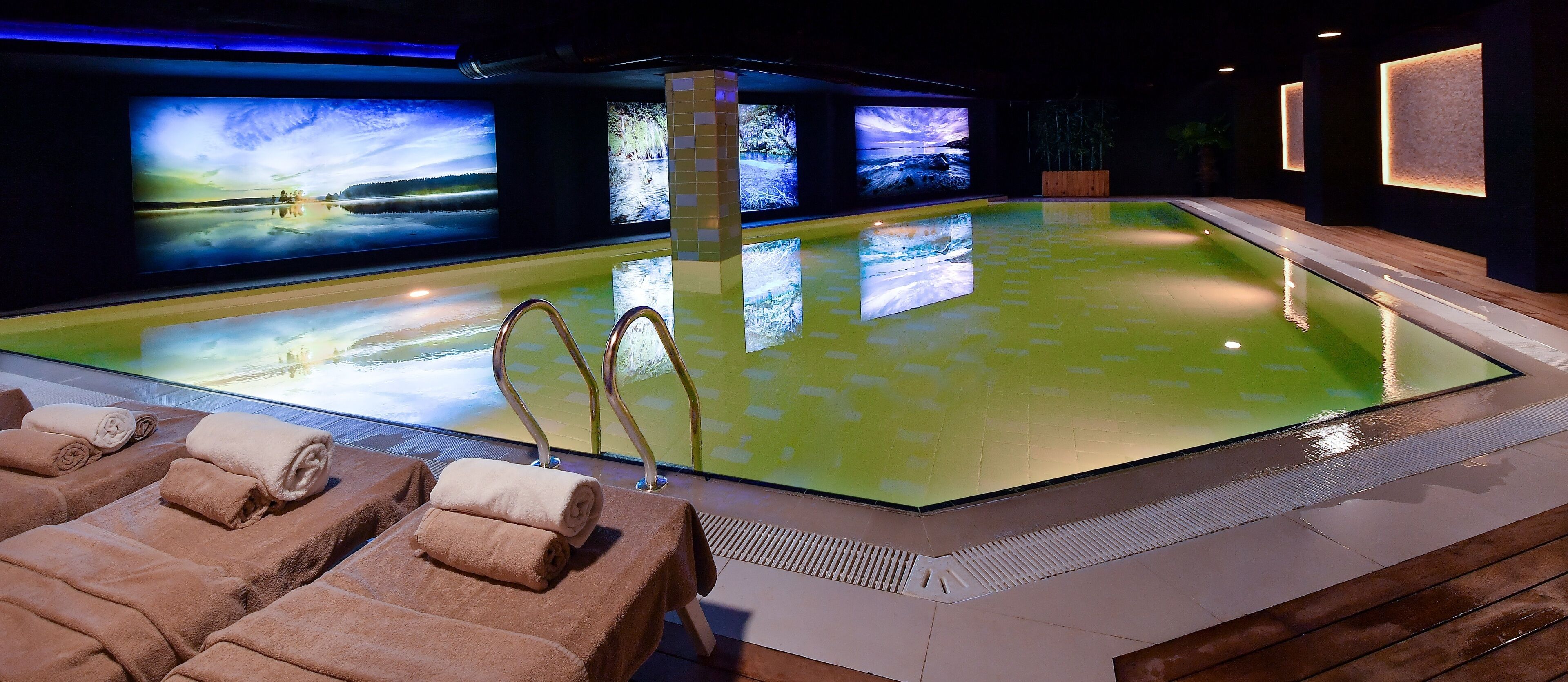 A spa area with a large pool, multiple screens displaying nature scenes, and several lounge chairs with towels.