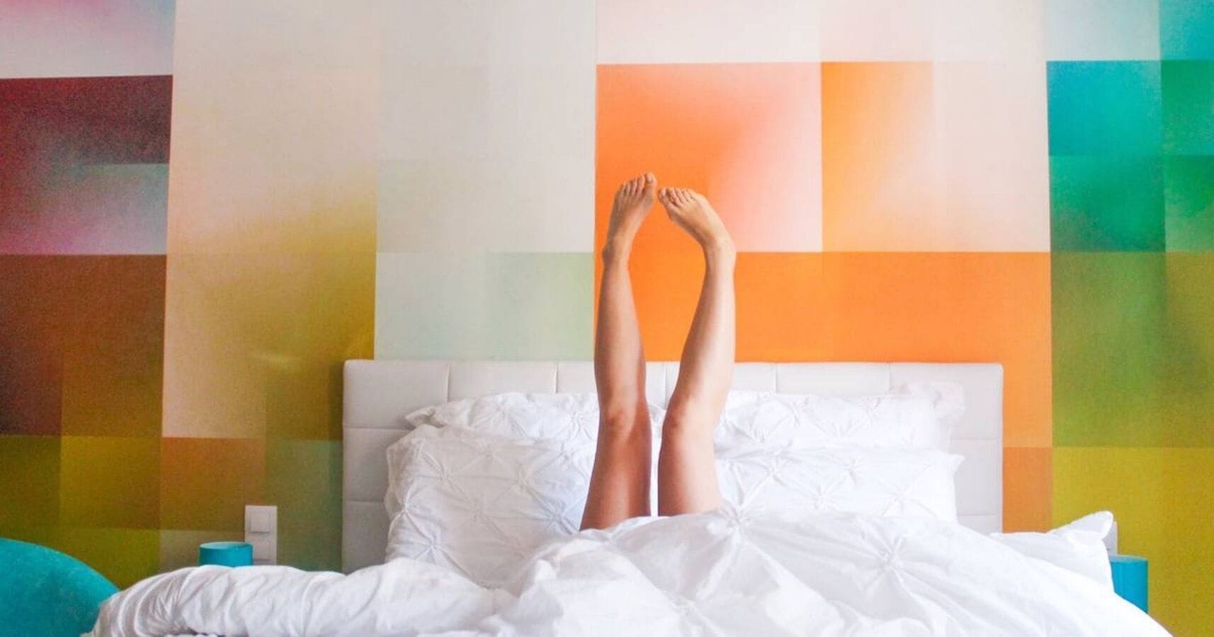 A bed with white bedding and a white headboard, set against a wall with a colorful, geometric pattern.