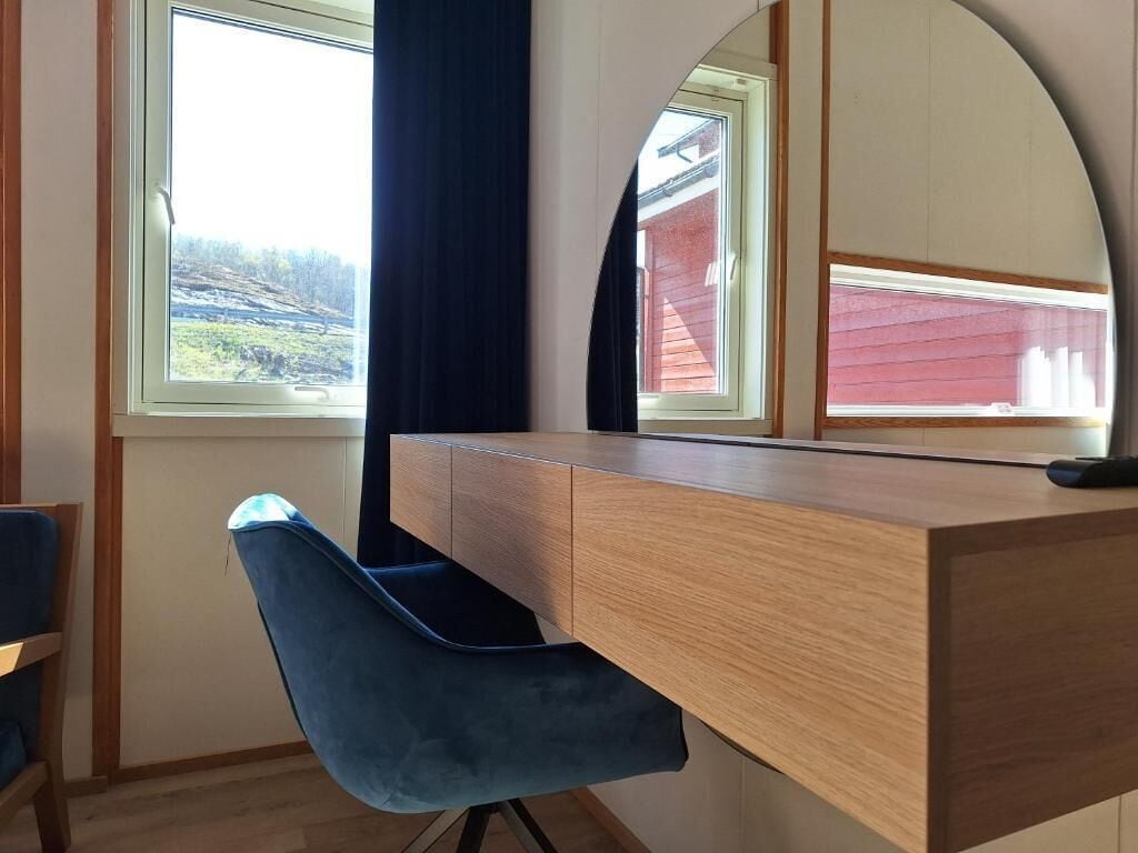 A room with a wooden desk, a blue chair, a window with a view of a red building, and a mirror reflecting the window.
