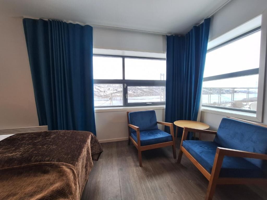 Habitación de hotel con una cama, dos sillones azules, una mesita redonda y ventanas amplias con cortinas.