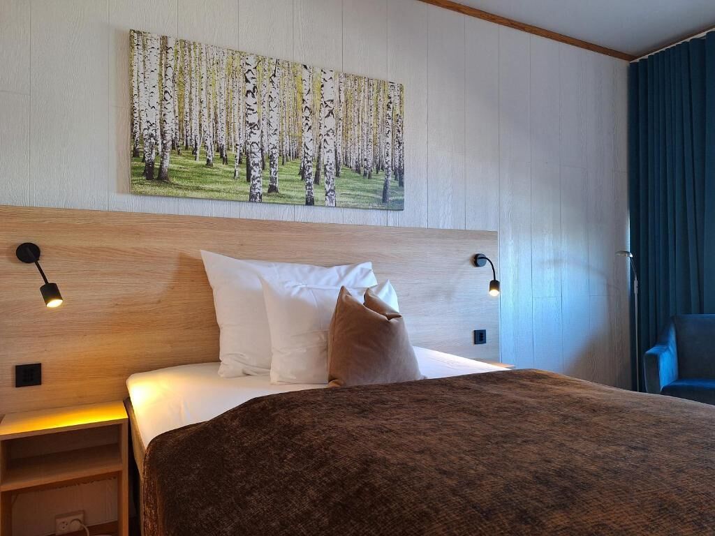 Ein Hotelzimmer mit einem großen Bett, zwei Nachttischlampen, einem Kopfteil, einem Waldgemälde und Blick ins Freie.