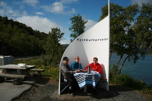 Three people seated at a table under a white tent with the text 'Kystkulturveien' on it, set against a backdrop of a lake and trees. - Tjeldsundbrua Hotel (Tjeldsund)