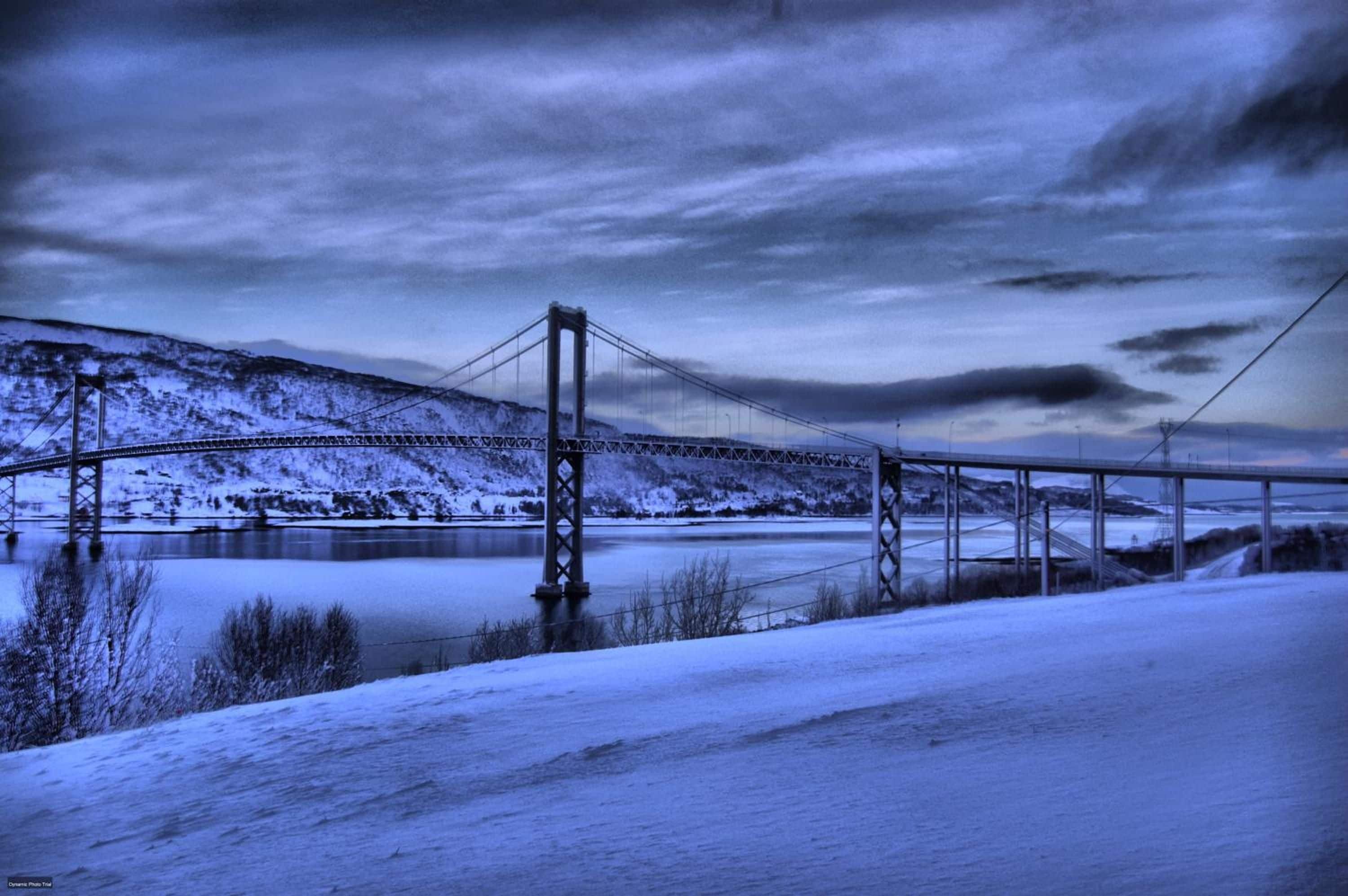 Een hangbrug over een bevroren rivier, met sneeuw bedekte oevers en heuvels op de achtergrond.