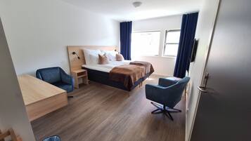 A hotel room with a bed, two chairs, a desk, and a trash bin.