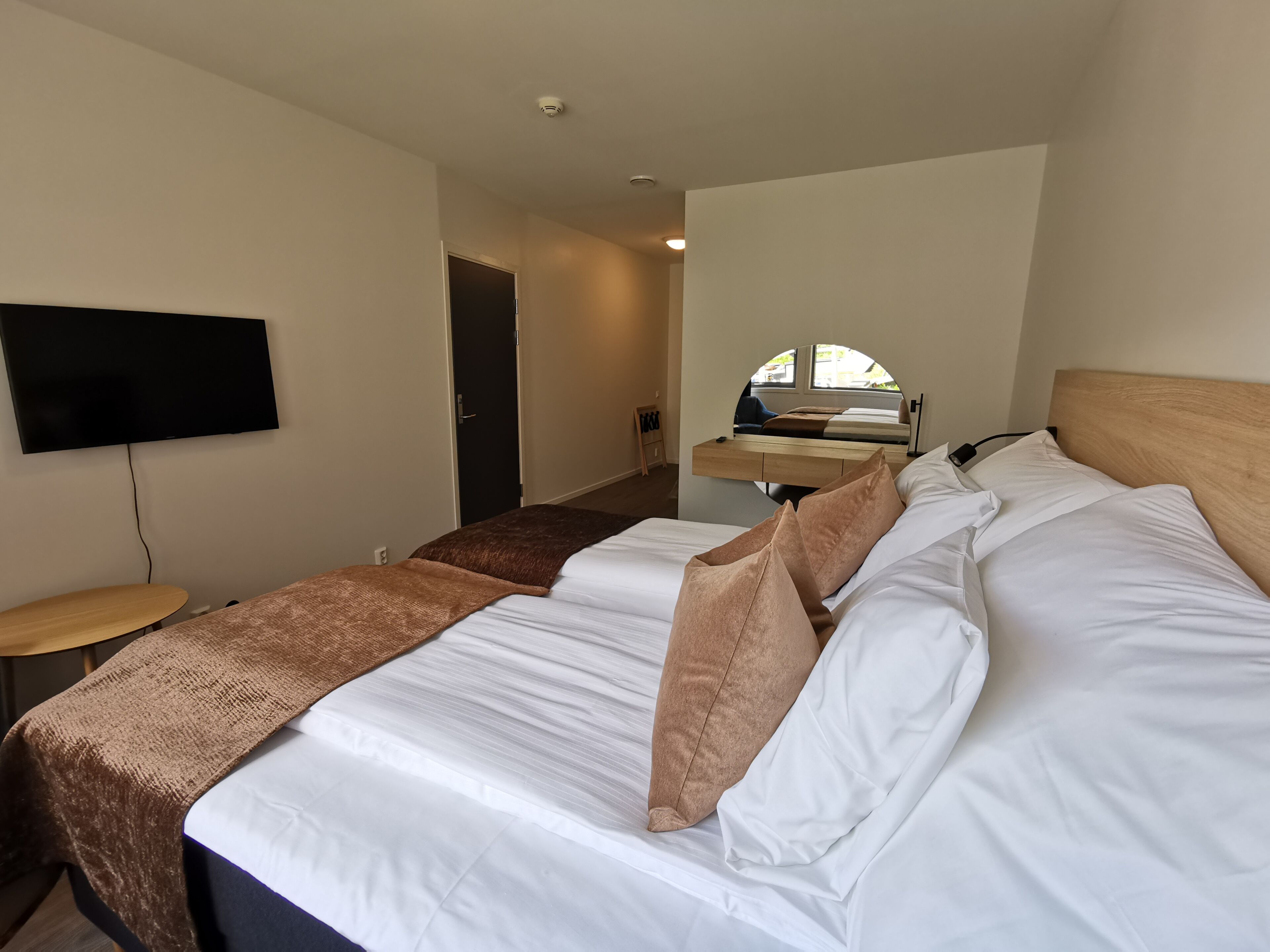 A hotel room with a bed, a TV mounted on the wall, a desk with a chair, and a window.