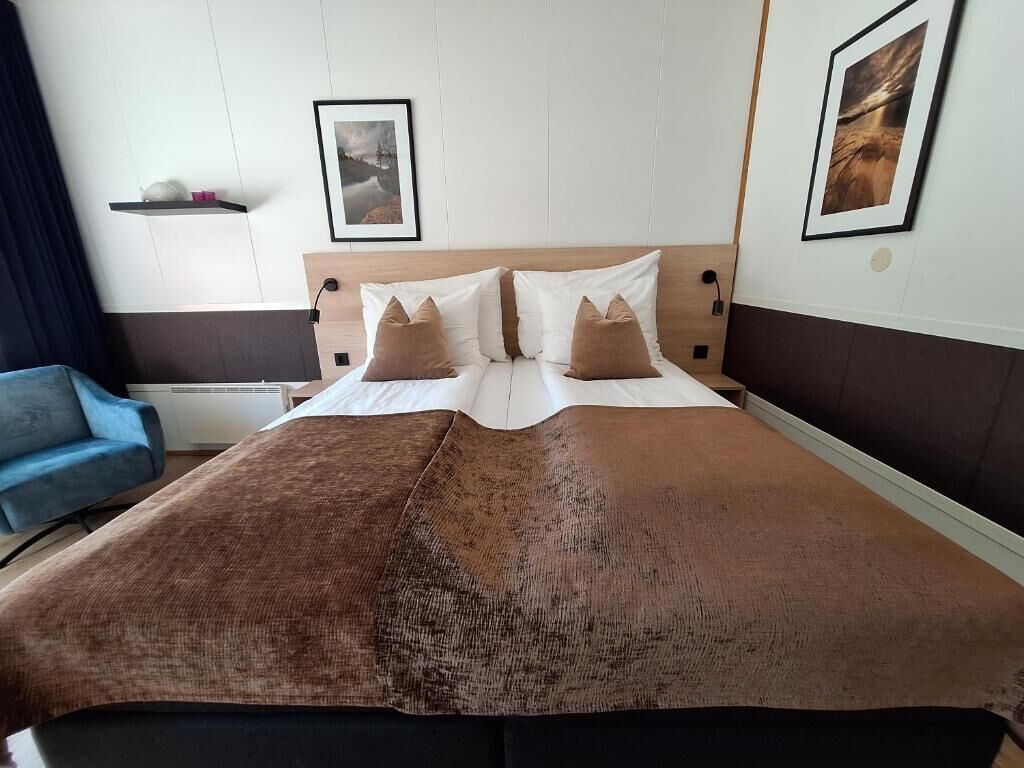 A neatly made bed with a brown blanket and pillows, a bedside table with a lamp, and a framed picture on the wall.