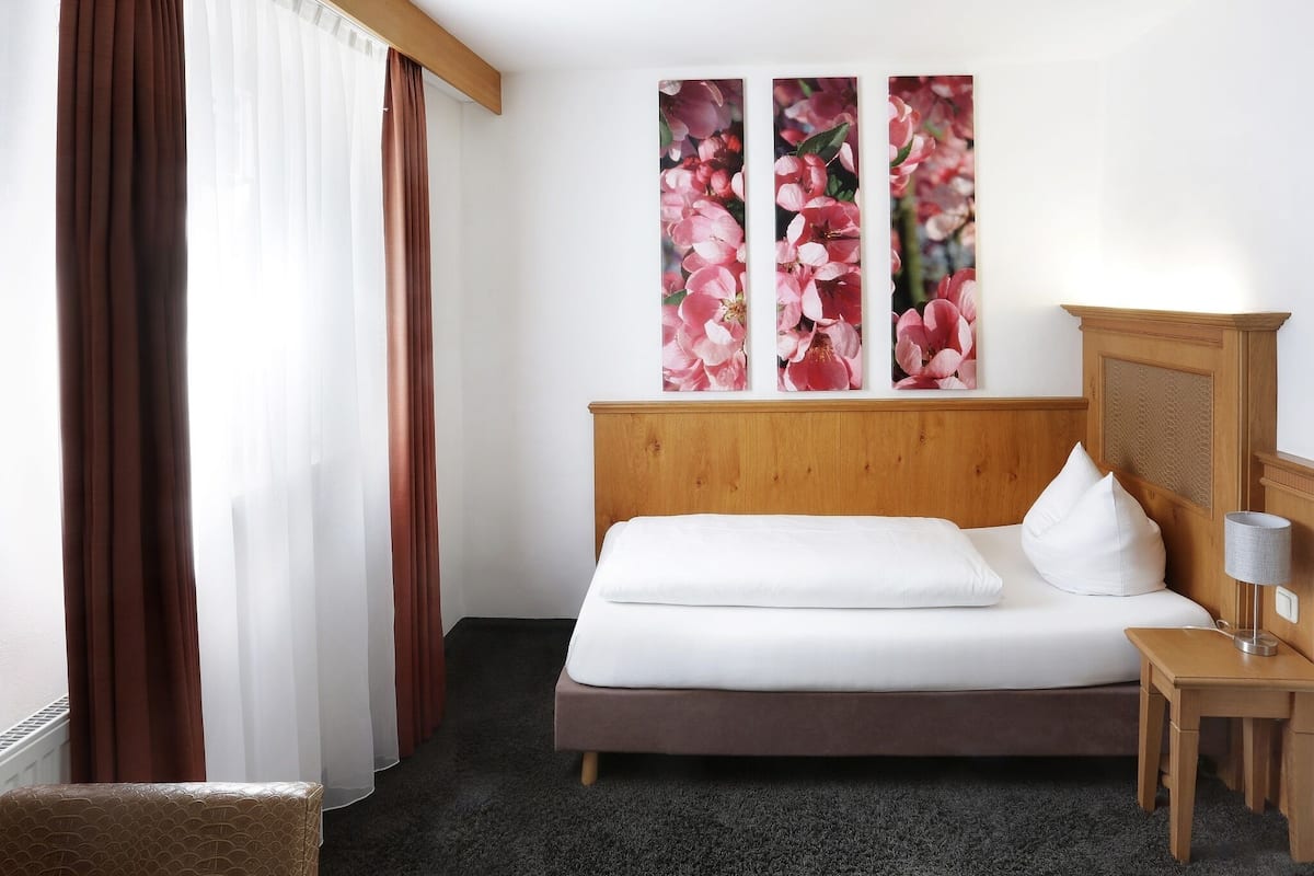 A hotel room with a wooden headboard, a bed with white linens, a small bedside table with a lamp, and a large floral artwork on the wall.