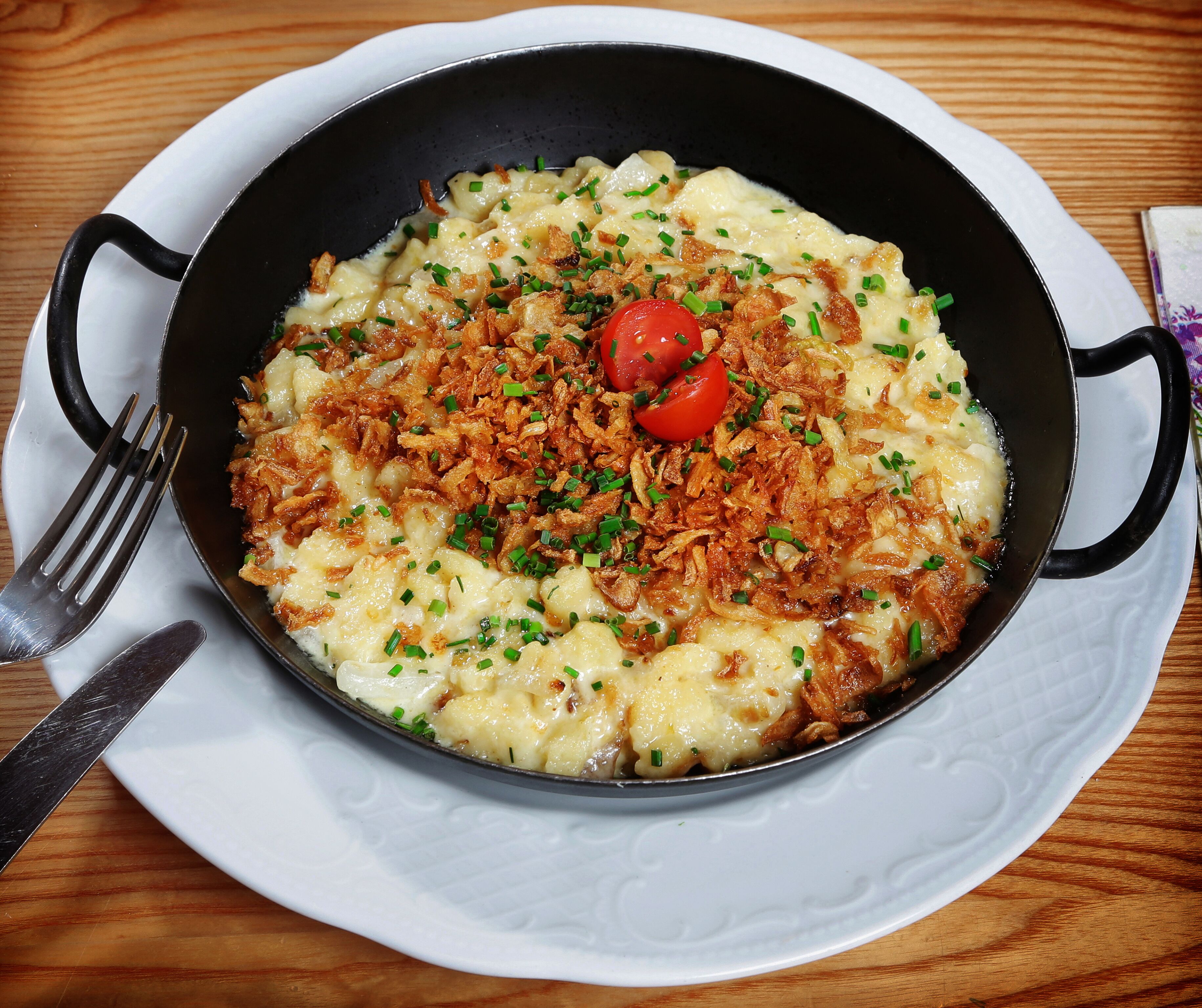 Una sartén con un plato de comida adornado con cebollín y un tomate cherry, servido en un plato blanco con un tenedor y un cuchillo a un lado.