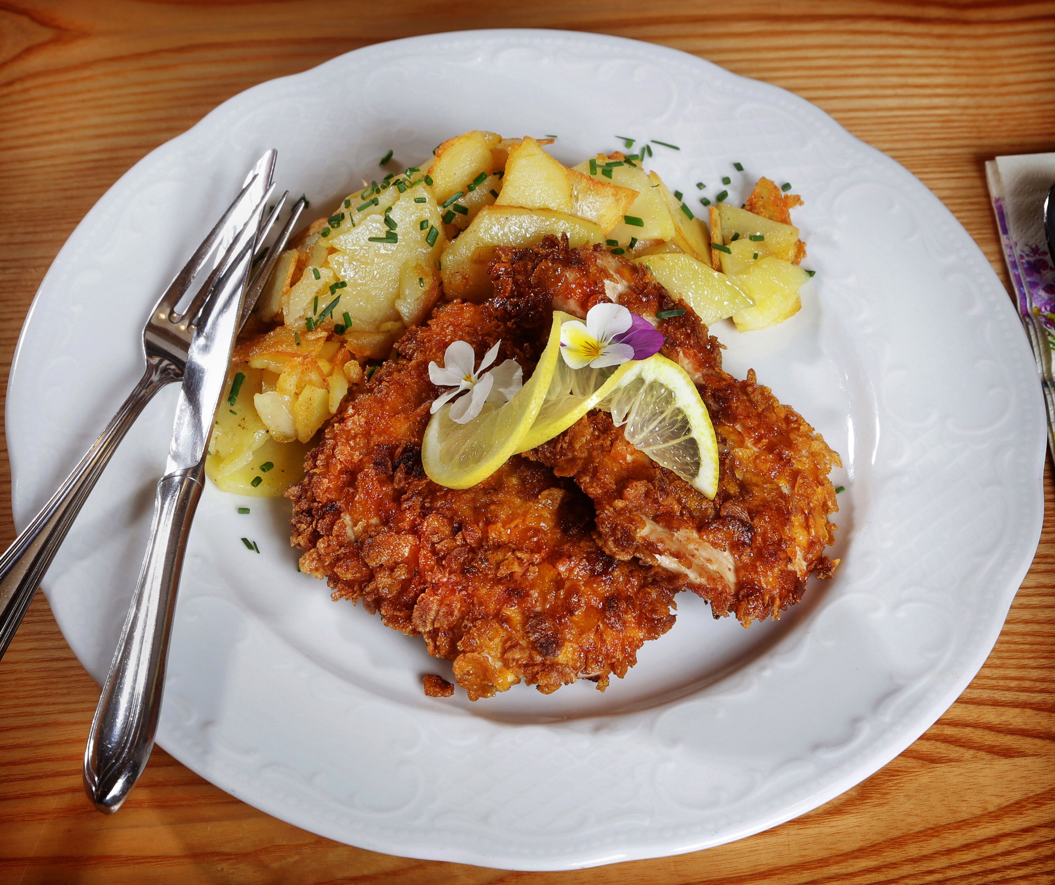 Un plato con una escalope empanizada y frita, decorado con limón y cebollín, acompañado de patatas hervidas y un tenedor sobre una mesa de madera.