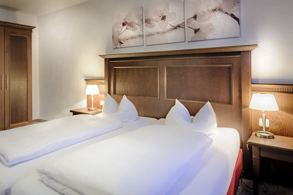 A hotel room with two beds, wooden headboard, white linens, two bedside lamps, and artwork on the wall.