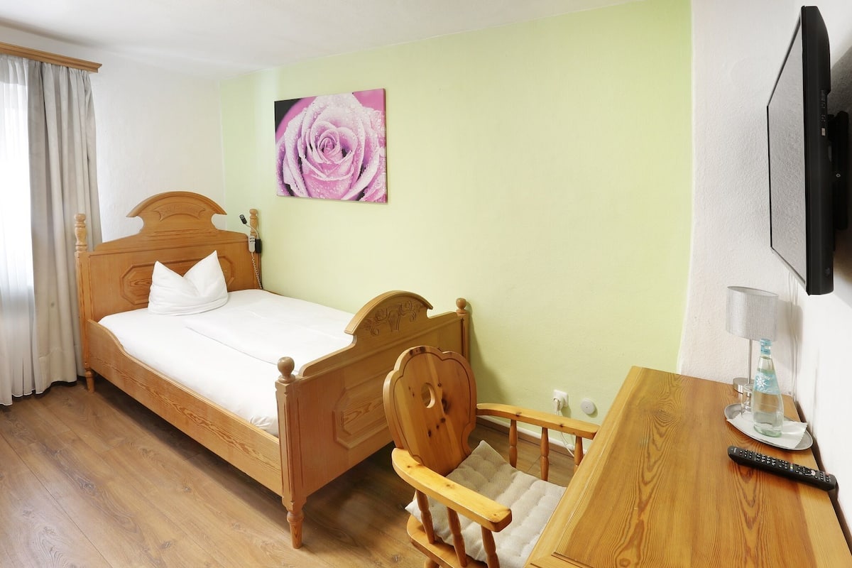 A hotel room with a wooden bed, a chair, a table, a TV, and a painting on the wall.