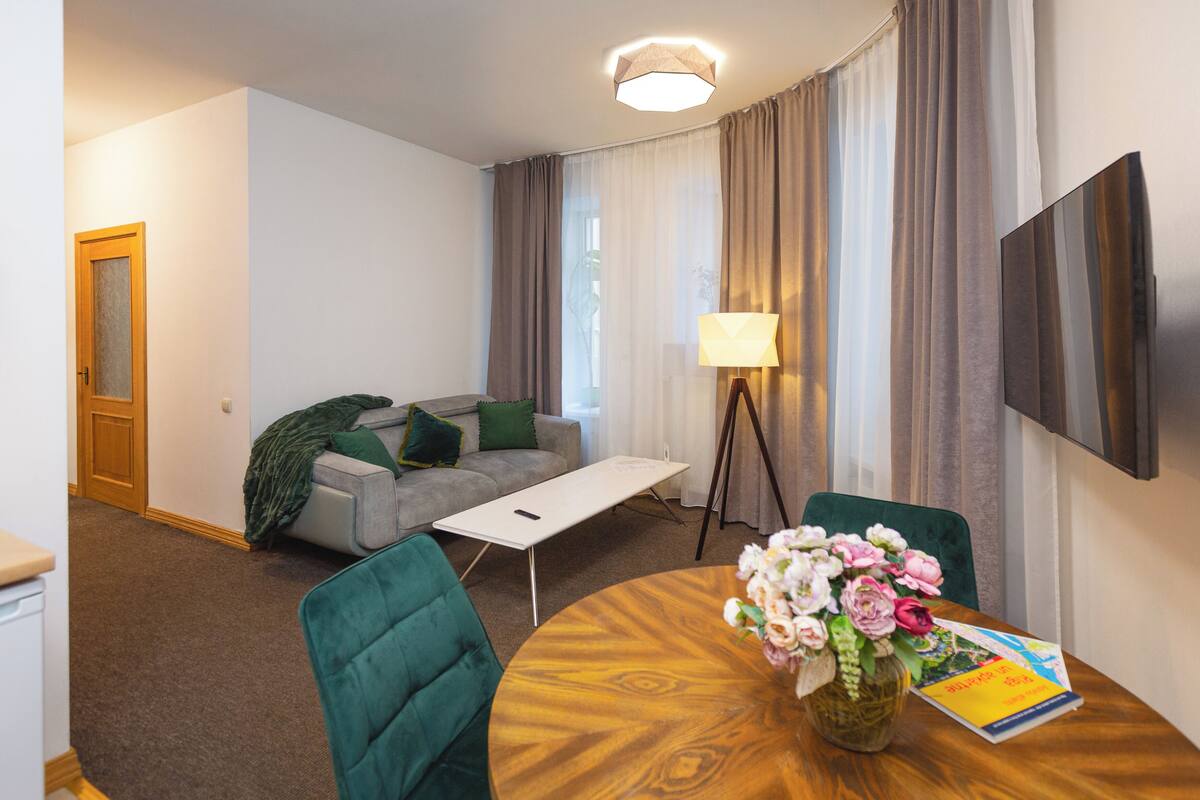 A modern living room with a sofa, a coffee table, and a dining table with chairs. There is a lamp, a flat-screen TV mounted on the wall, and a door.