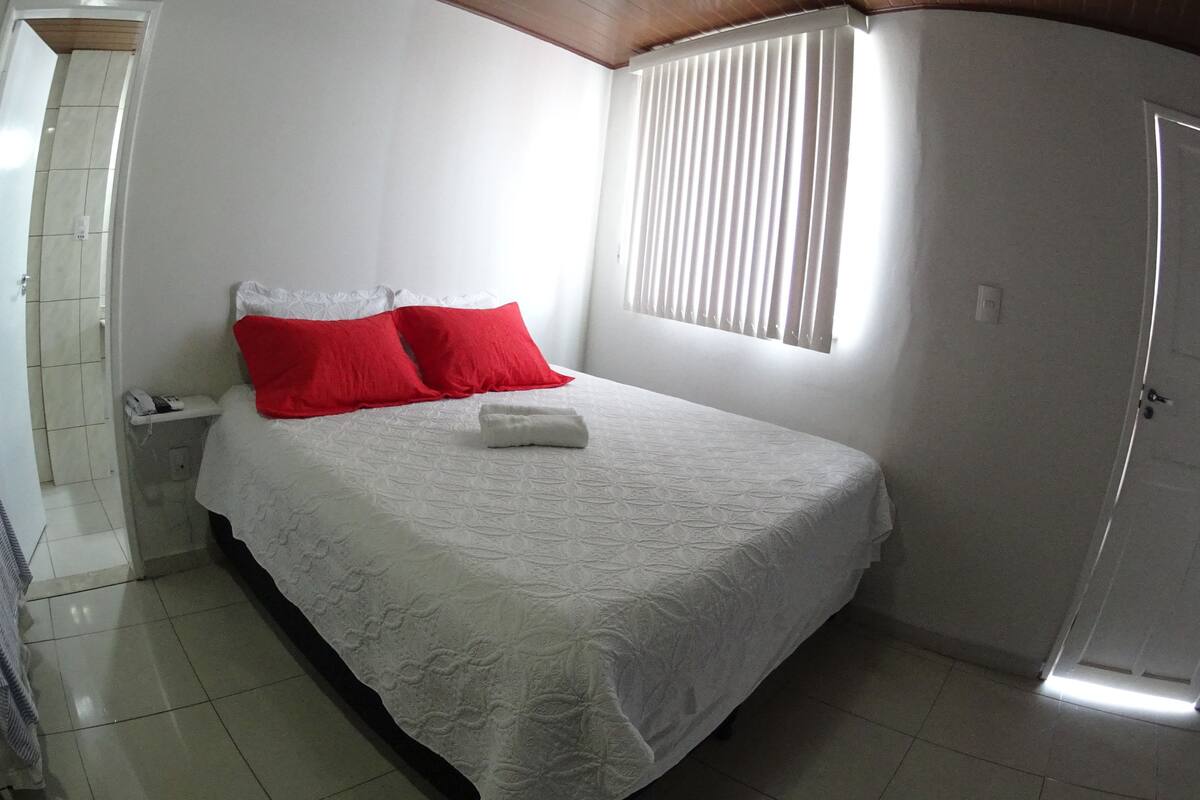 A neatly made bed with white bedding and red pillows, a bedside telephone, and a window with blinds.