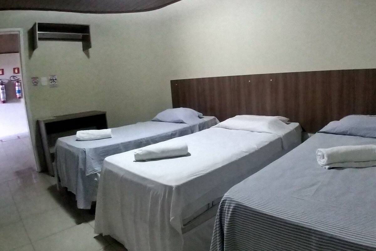 A hotel room with three single beds, each with a white bedsheet and pillow, and a wooden headboard.