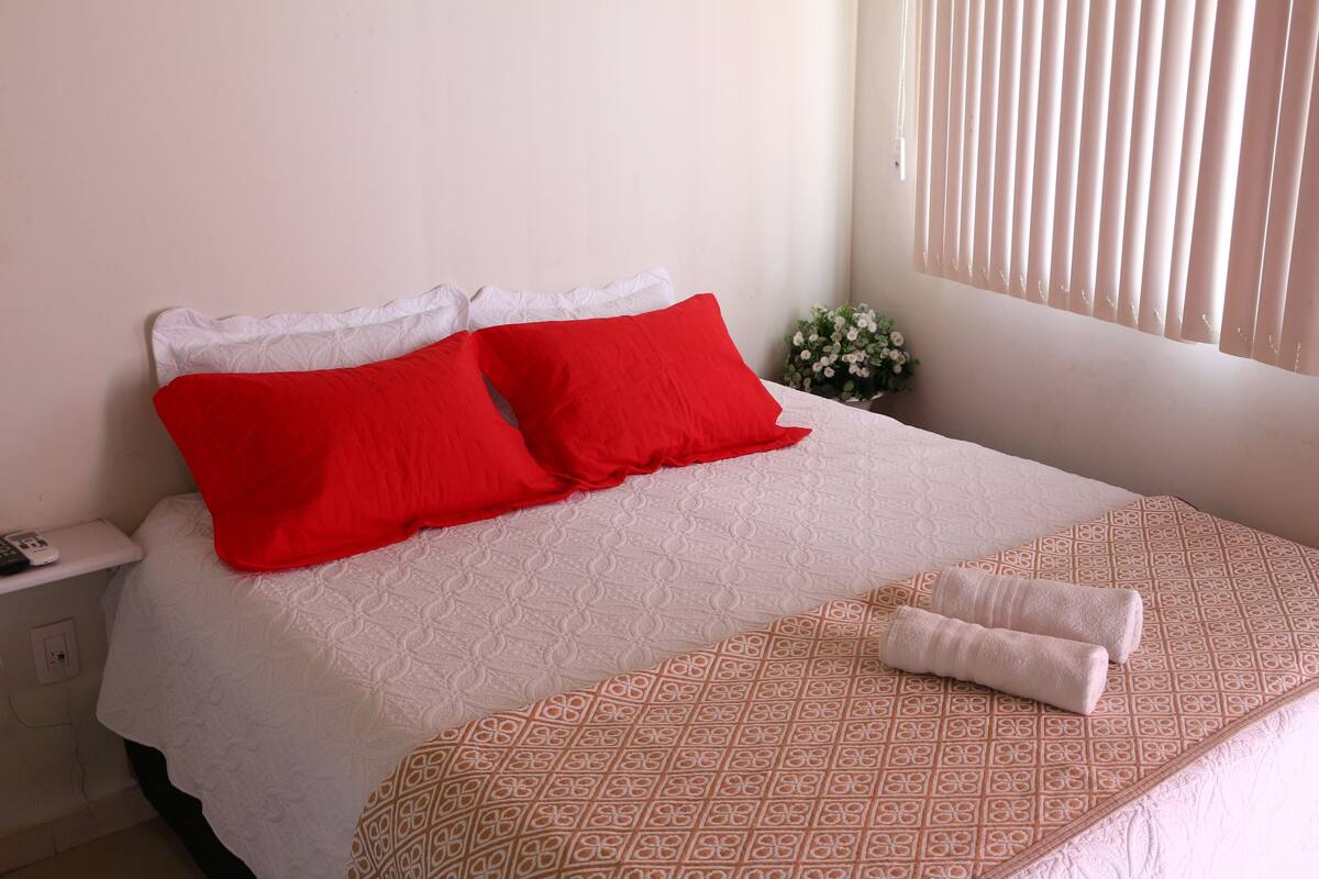 A neatly made bed with a white and patterned bedspread, two red pillows, and a folded towel on the side.