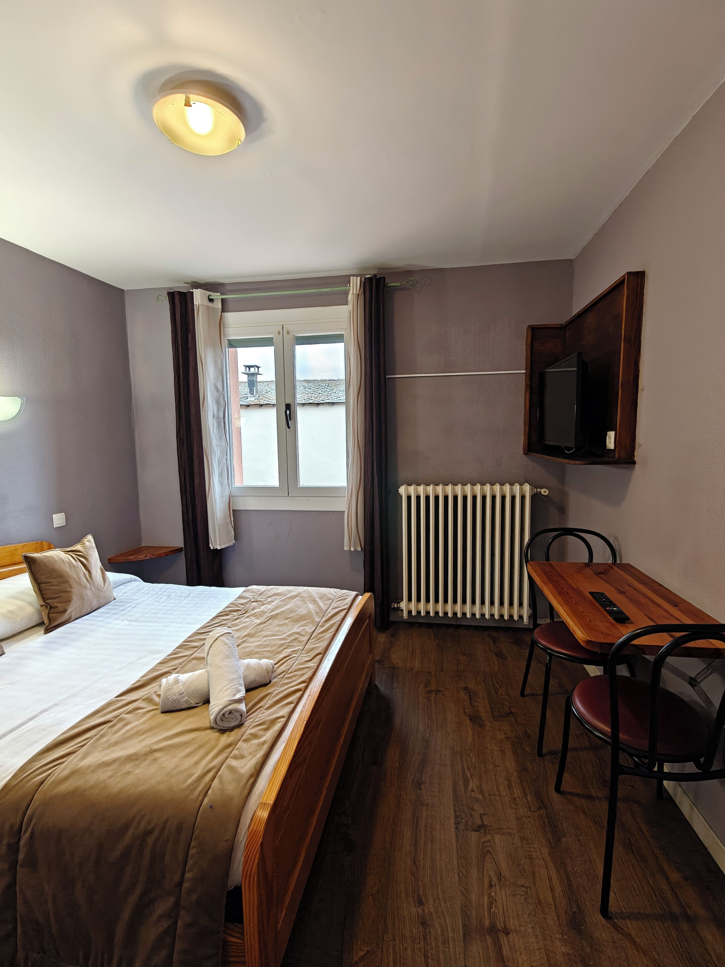 Una camera d'albergo con un letto, un tavolo in legno con due sedie, una televisione a parete e una finestra con tende.