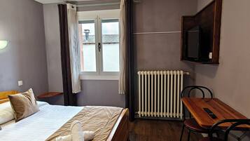 Una habitación de hotel con cama, una mesa de madera con dos sillas, un televisor montado en la pared y un ventanal con cortinas.