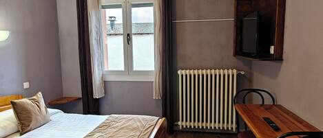 Una habitación de hotel con cama, una mesa de madera con dos sillas, un televisor montado en la pared y un ventanal con cortinas.