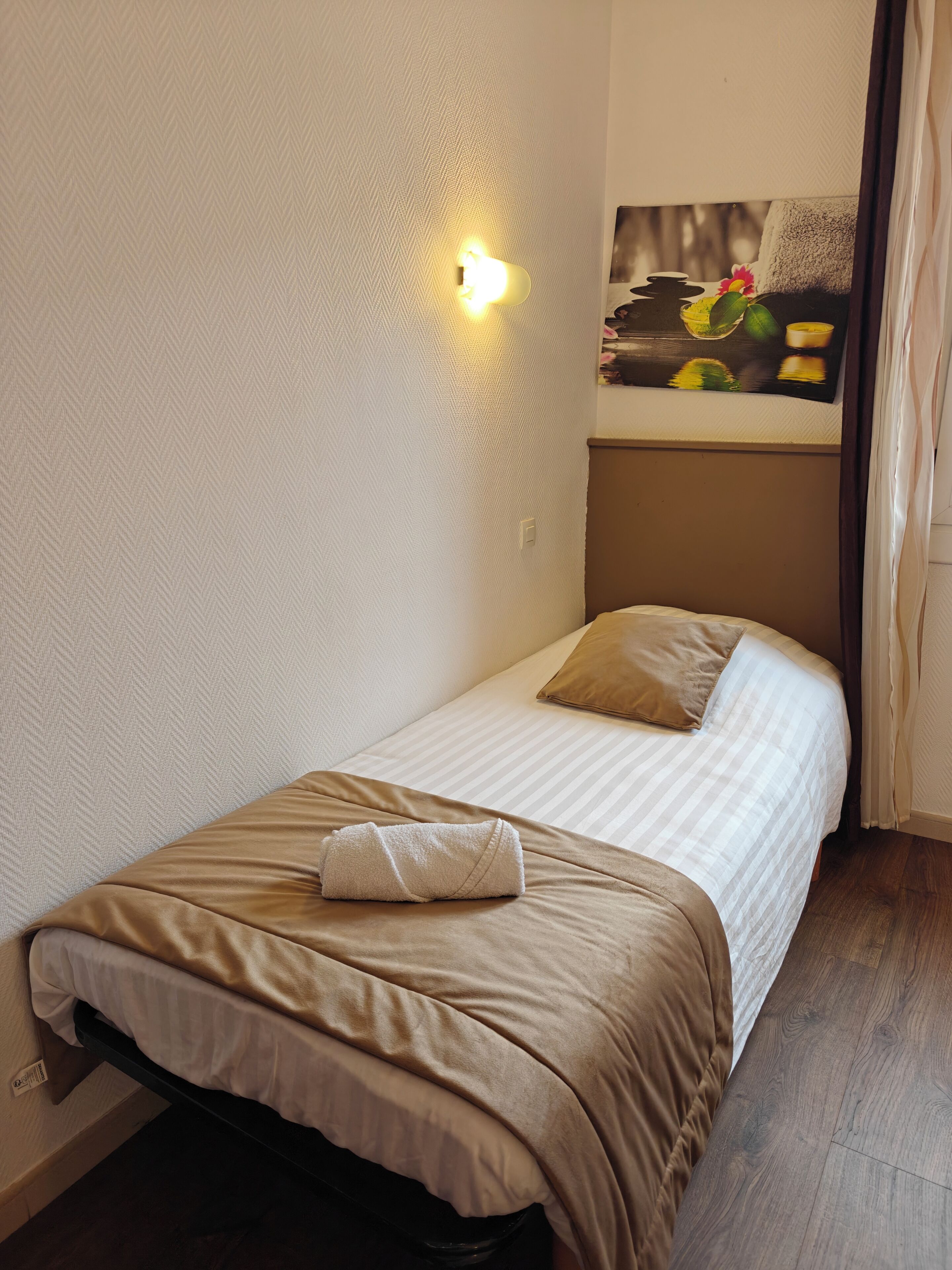 Een enkel bed met een beige dekbedovertrek en kussen, een klein nachtkastje met een lamp, en een schilderij boven het bed.