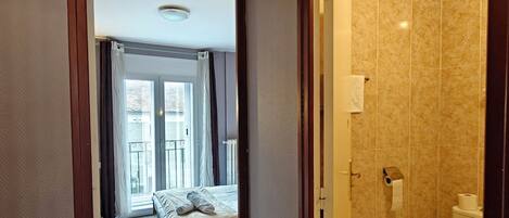 Habitación de hotel con cama, baño con inodoro y ducha, y pasillo que da a un balcón.