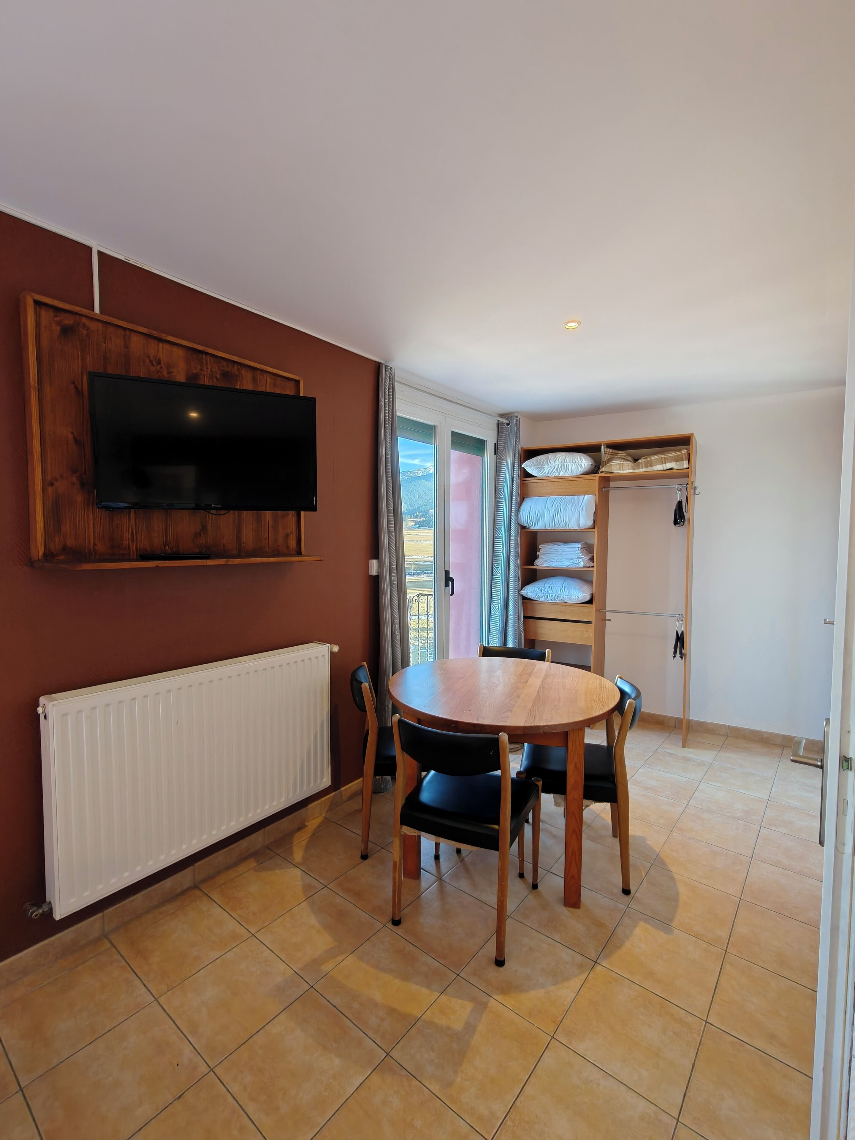 Un comedor con una mesa y sillas de madera, un televisor montado en la pared y vistas a través de un ventanal corredero.
