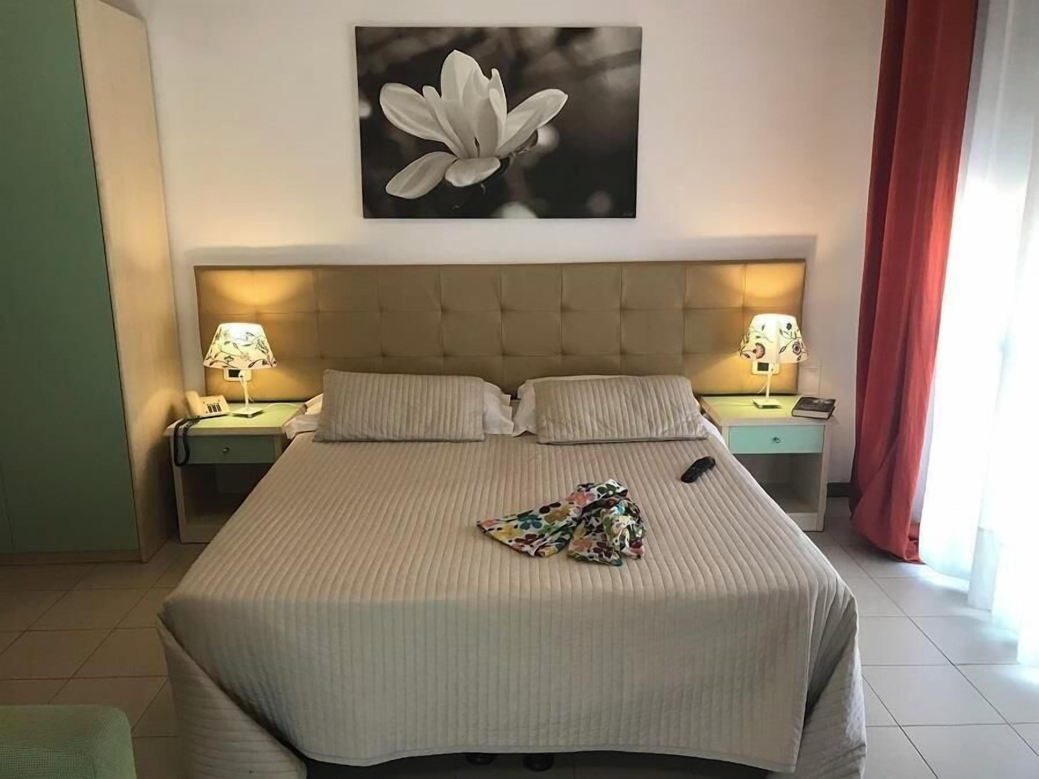 Ein Hotelzimmer mit einem großen Bett, zwei Nachttischen mit Lampen, einem Blumenbild an der Wand und einem Fenster mit roten Vorhängen.