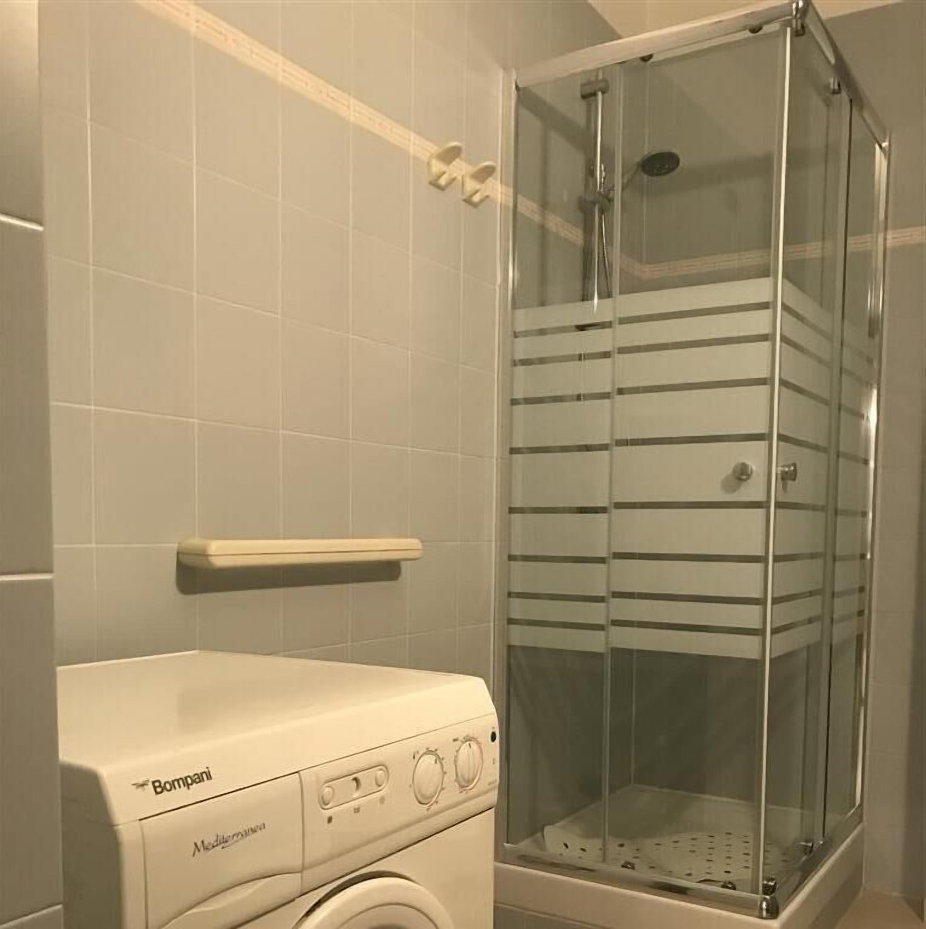 Un baño pequeño con lavadora, cabina de ducha de vidrio y pared de azulejos.