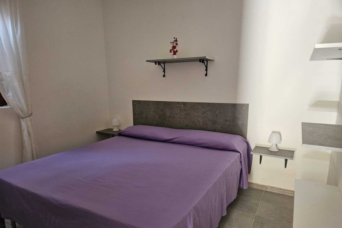 A bedroom with a bed, a nightstand, a wall-mounted shelf, and a bedside table.