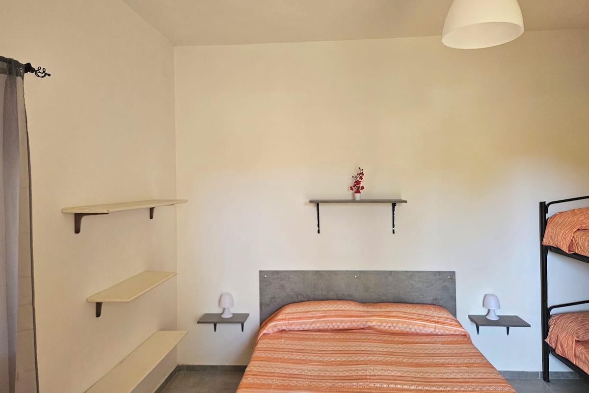 A bedroom with a bed, a bedside table, a hanging light, and a shelf with a plant.