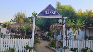 A sign for 'ULISSE LOUNGE BAR' with a pathway leading to a white picket fence and thatched-roof structures. - Camping Ulisse (Lamezia Terme)