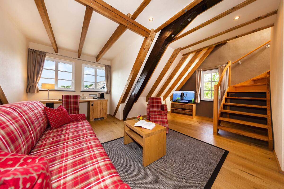 A cozy loft bedroom with a red plaid sofa, wooden beams, a wooden staircase, and a view of the outdoors.