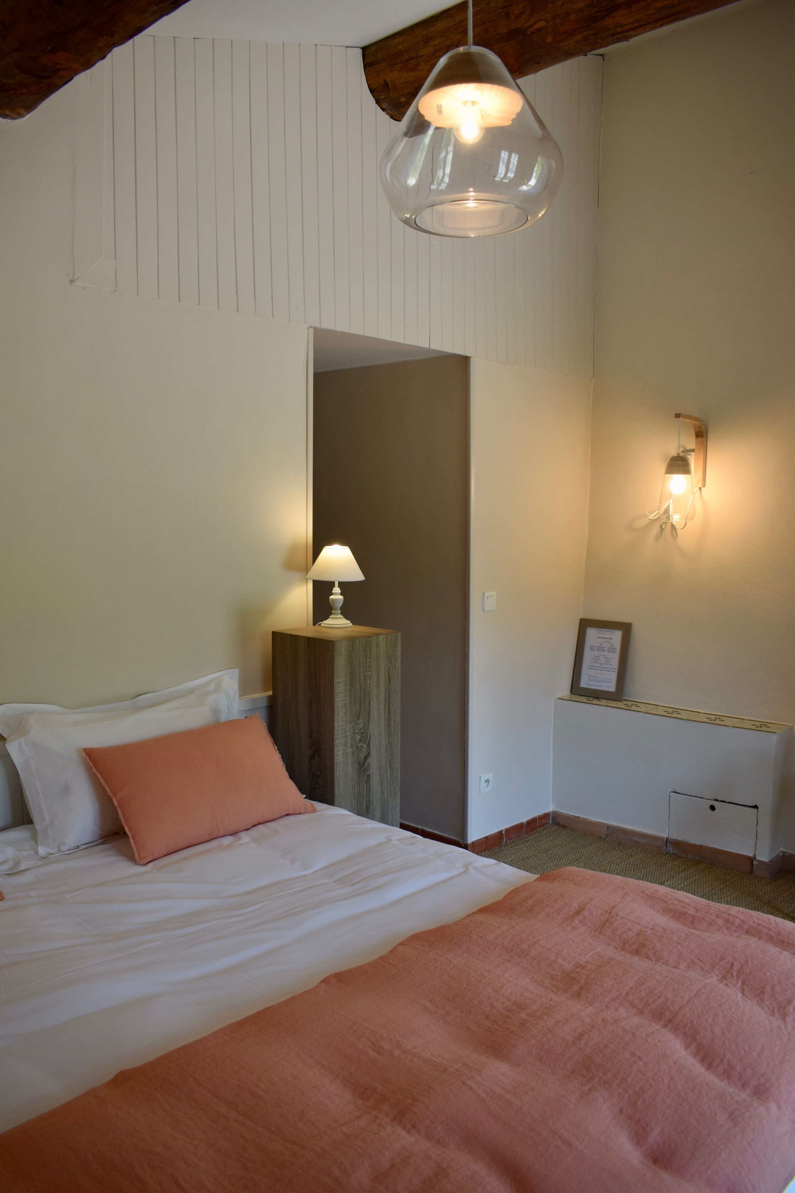 A bedroom with a bed, a bedside table with a lamp, a wall-mounted light, and a framed certificate on the wall.