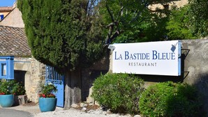 A sign for "La Bastide Bleue Restaurant" with a stone wall, blue door, and potted plants. - La Bastide Bleue (Seguret)