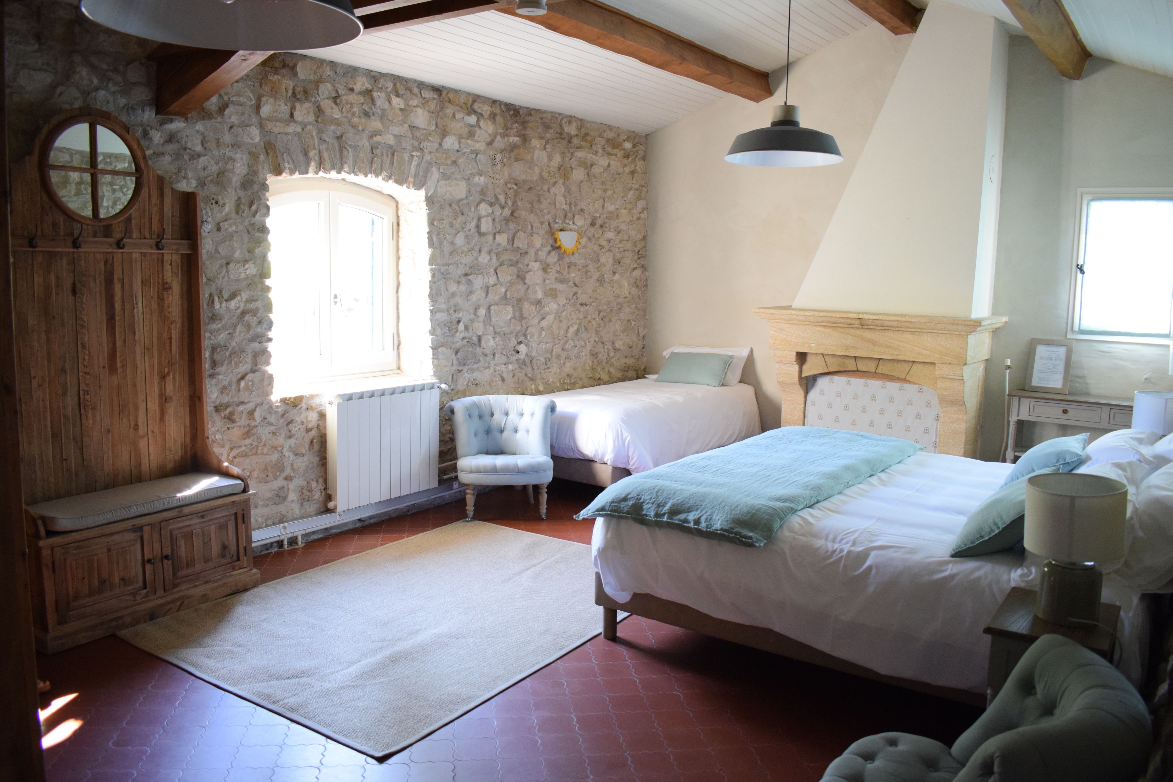 A bedroom with a stone wall, wooden furniture, a bed with a headboard, a bench, a chair, and a window.
