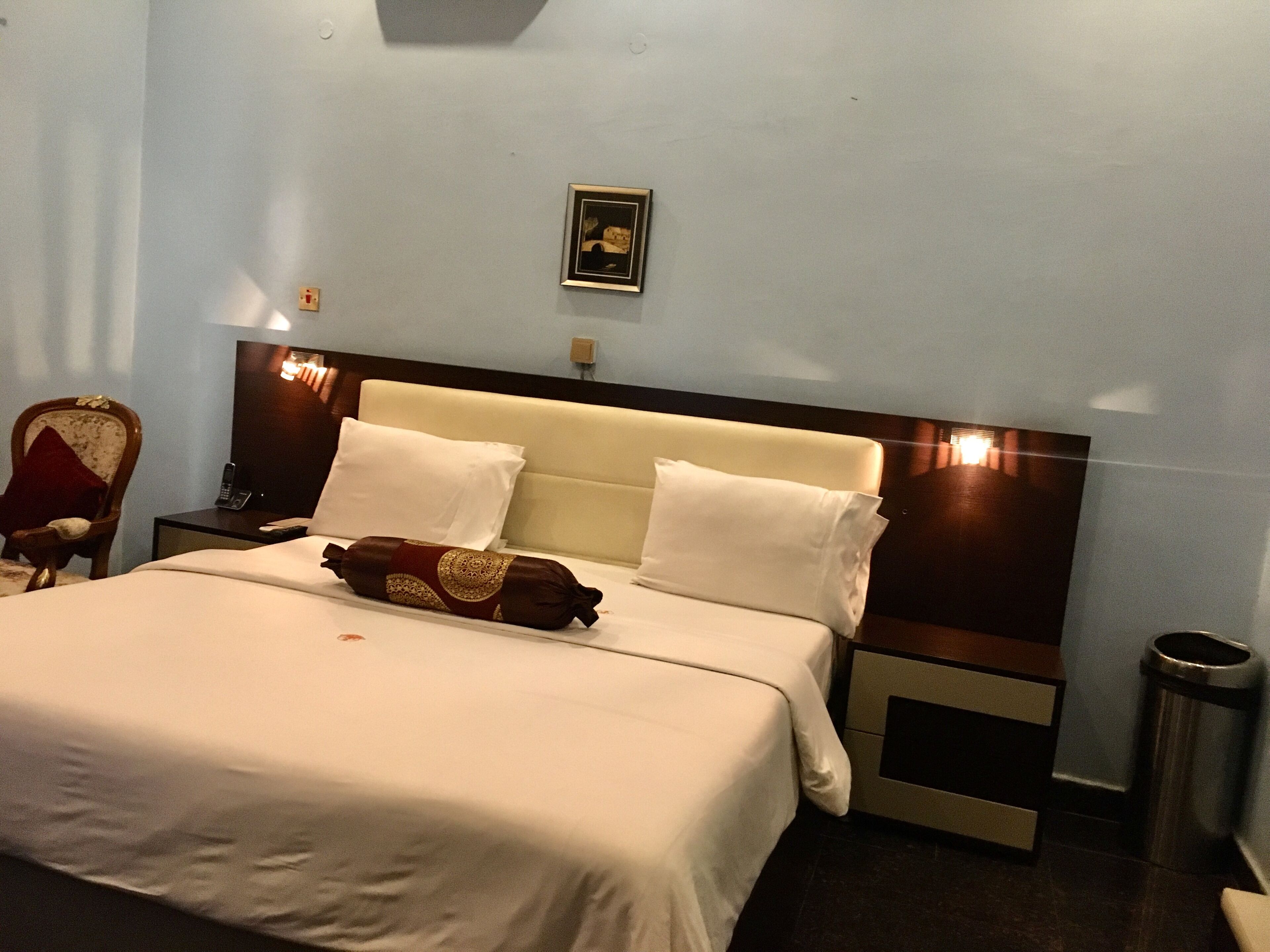 A hotel room with a bed, a nightstand, a chair, a picture on the wall, and a trash can.